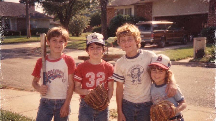 Four young boys stand together on a suburban street, with one holding a baseball bat and two wearing baseball gloves.