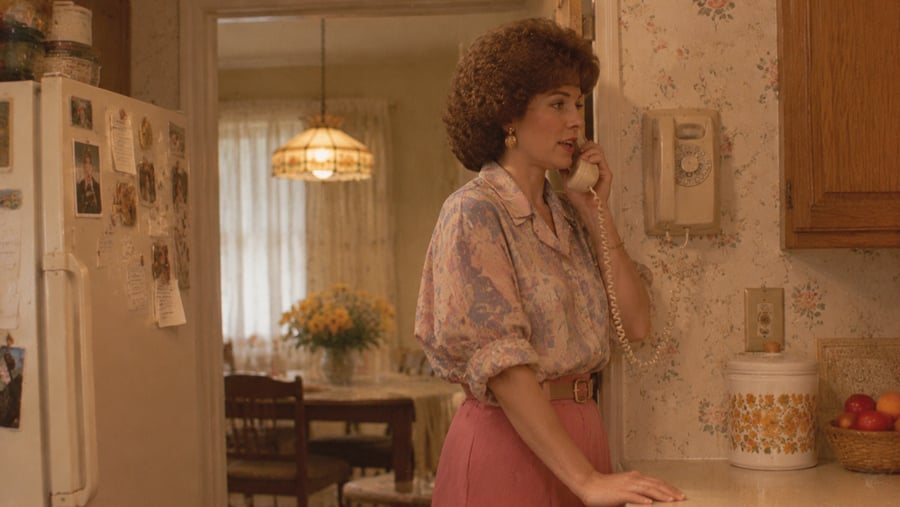 A woman with curly hair wearing a patterned blouse and pink skirt stands in a kitchen talking on a wall-mounted rotary phone.