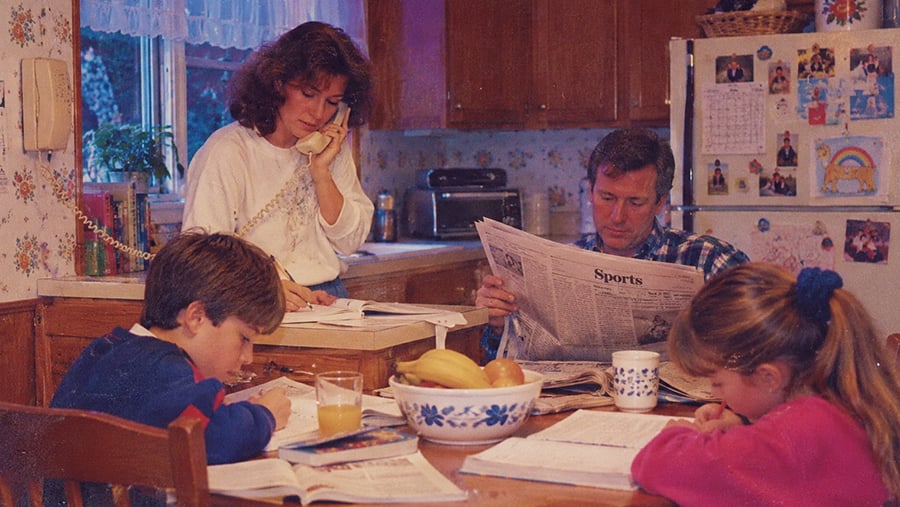 A family sits at a kitchen table; the mother talks on a wall-mounted phone while the father reads the sports section, and two children work on school assignments.