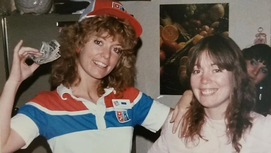 A Domino’s pizza employee wearing a company hat and uniform holds up a handful of cash while posing with another woman.