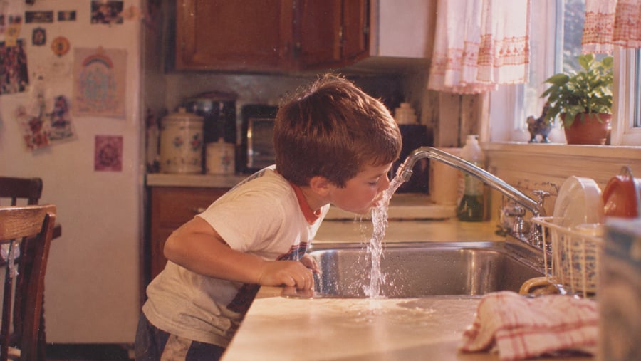 A young boy leans over a kitchen sink to drink water directly from the faucet.
