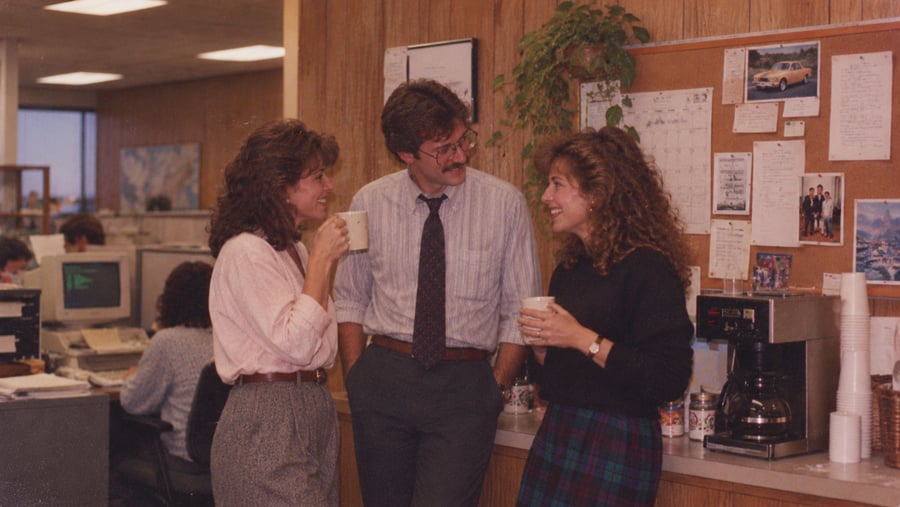 Two women and a man stand in an office break area, talking and holding coffee mugs.