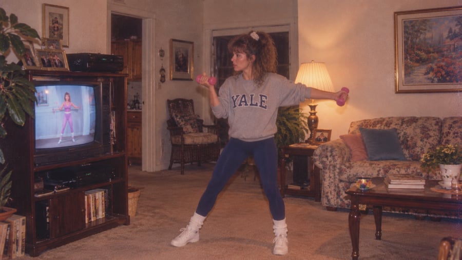 A woman in a Yale sweatshirt and leggings exercises with hand weights while following a workout video on television.