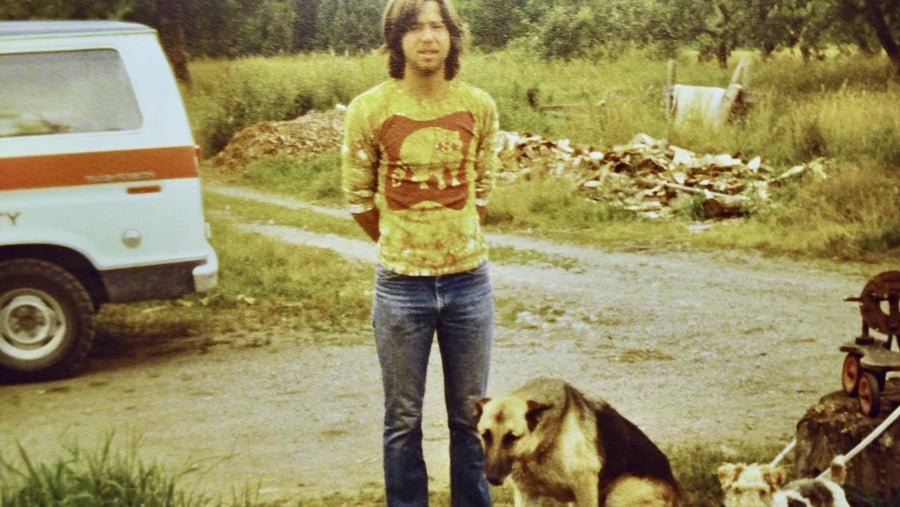 A man wearing a patterned yellow shirt and jeans stands on a gravel path next to a van and two dogs.