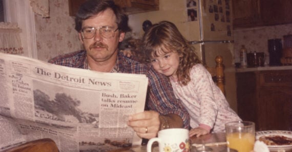 A man with glasses and a mustache reads The Detroit News while a young girl leans against his shoulder.