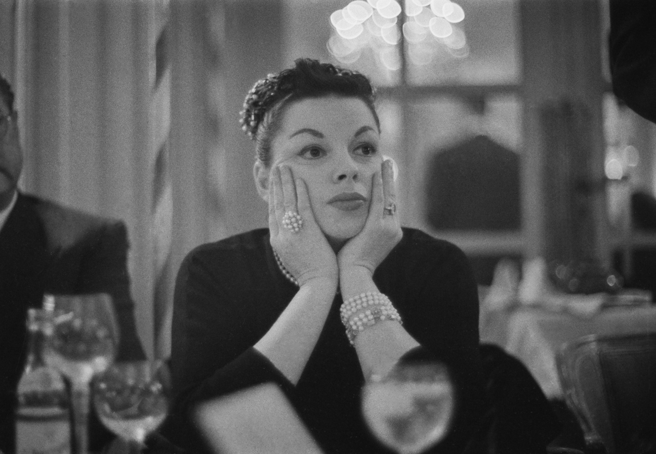Judy Garland resting her chin on her hands, wearing several pearl bracelets and a large ring.