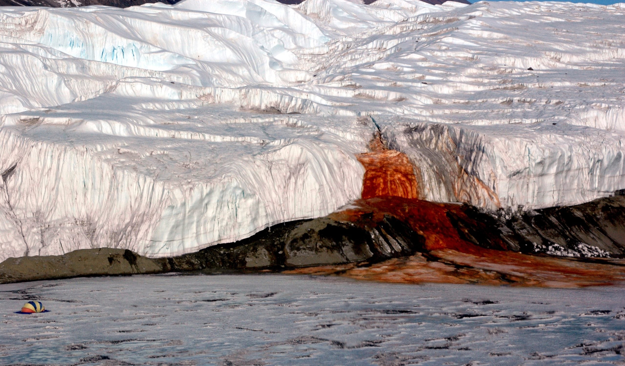 A glacier with a streak of bright red, iron-rich water flowing from it onto a frozen lake.