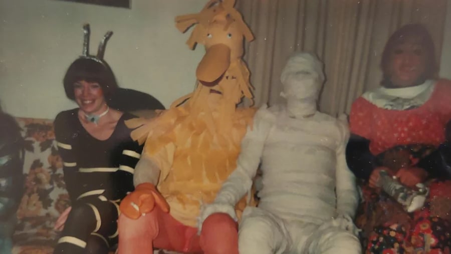 Four people in costumes—a bee, a large bird-like creature, a mummy, and a person in a patterned dress—sitting on a sofa.