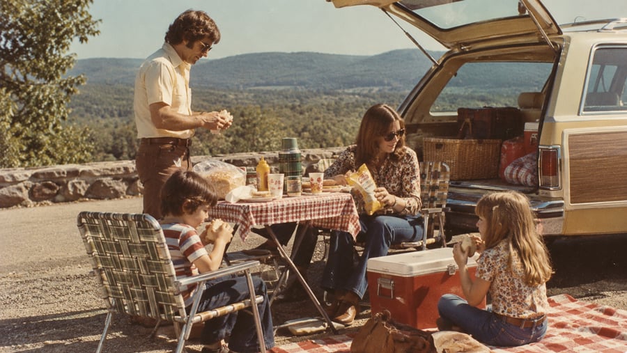 A family has a picnic at a scenic overlook next to their station wagon.