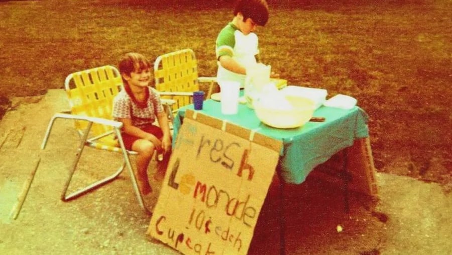 Two children run a lemonade and cupcake stand with a hand-lettered sign on a table outdoors.