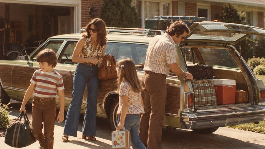 A family of four prepares to load luggage into the back of a vintage station wagon in a residential driveway.