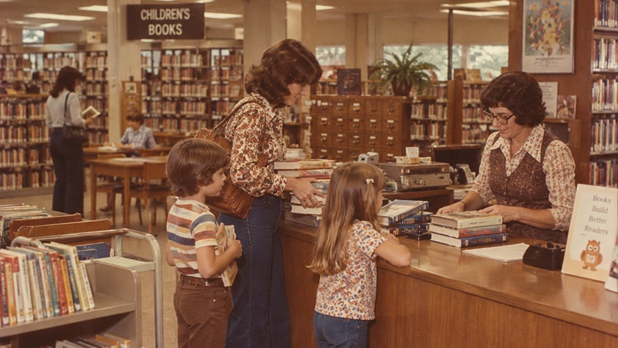 A librarian processes books for a woman and two children at a library circulation desk.