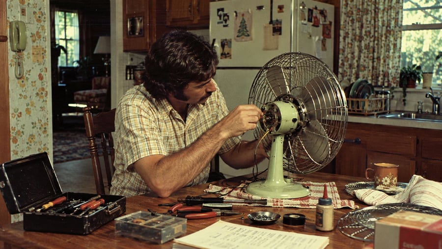A man in a plaid shirt repairs an electric desk fan while sitting at a kitchen table surrounded by tools.