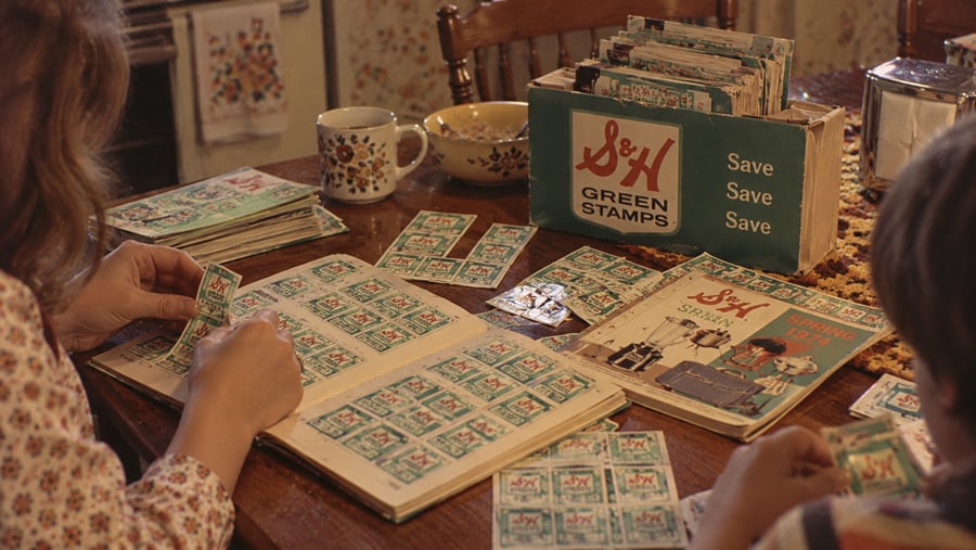 Two people sit at a wooden table pasting S&H Green Stamps into collection booklets, with a box of stamps and a catalog nearby.