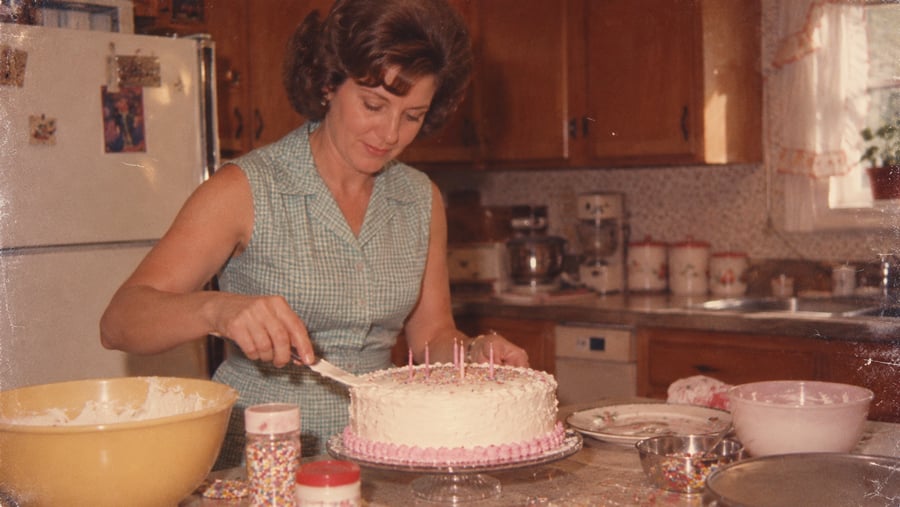 A woman in a gingham dress frosts a birthday cake with candles in a kitchen.