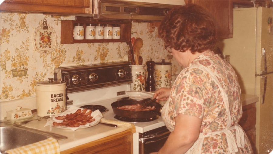 A woman in a floral dress and apron cooks bacon in a skillet on a stove next to a plate of cooked bacon and a container labeled “Bacon Grease.”