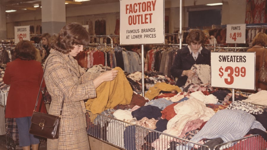Shoppers browse through bins of clothing in a store with signs for “Factory Outlet,” “Dresses,” “Pants,” and “Sweaters” priced at $3.99.