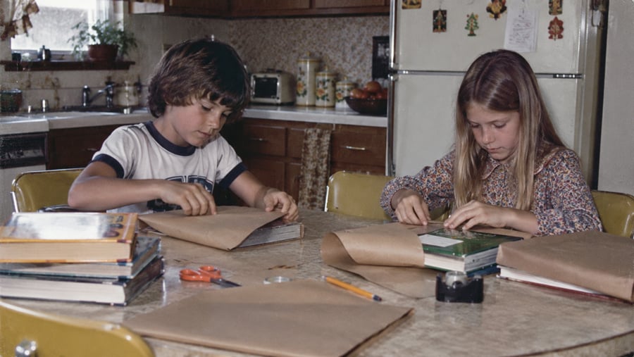 A boy and girl sit at a kitchen table wrapping school textbooks in brown paper.