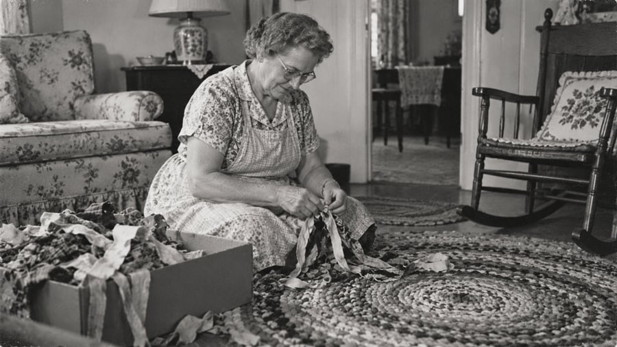 An older woman wearing glasses and an apron sits on a rug, braiding fabric strips from a cardboard box.