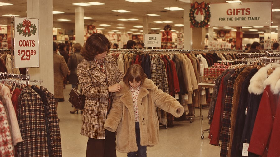 A woman helps a young girl try on a tan faux-fur coat in a clothing store with holiday signage for coats and gifts.