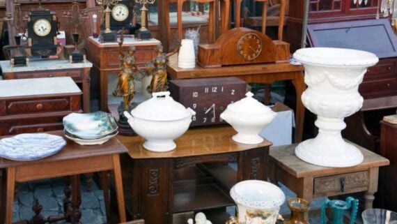 An assortment of antique clocks, two white lidded tureens, and a large white pedestal urn displayed on wooden tables at an outdoor market.