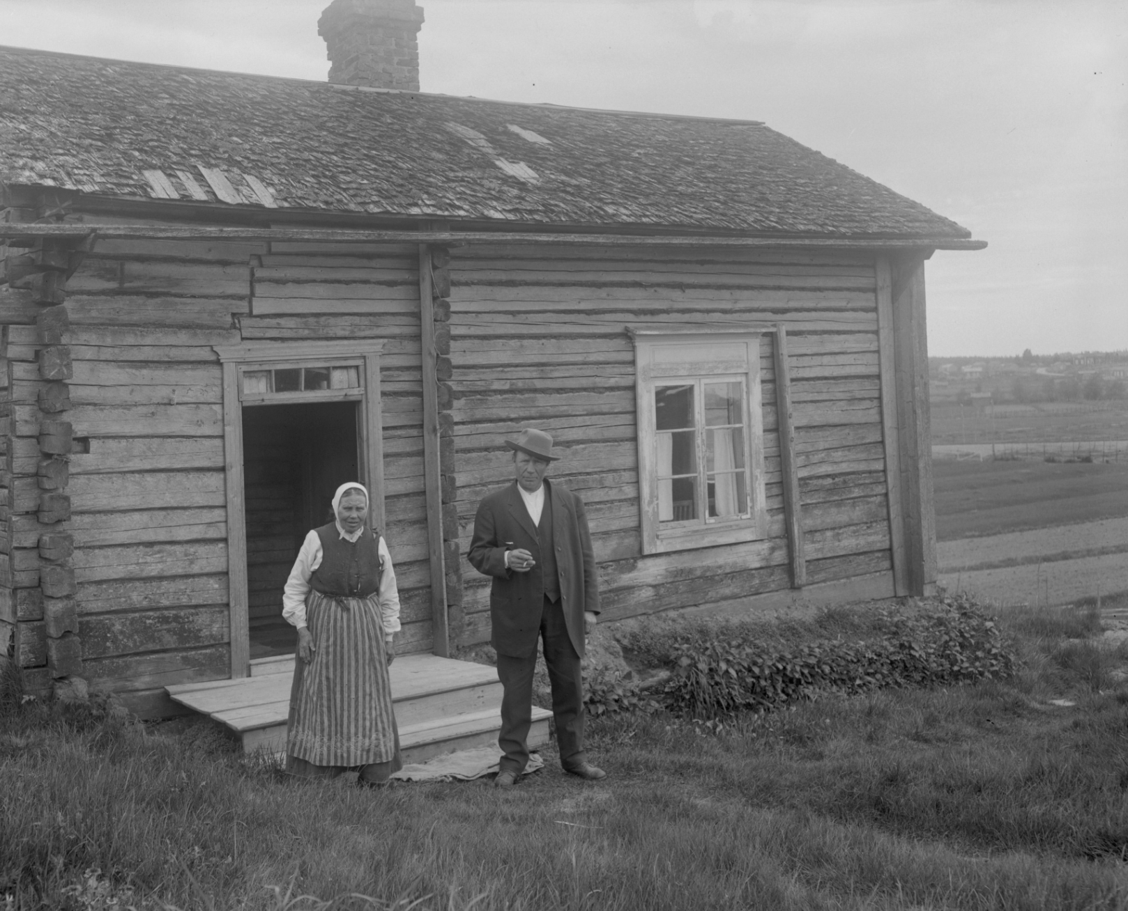 A man in a suit and hat stands next to a woman in an apron in front of a rustic log cabin.
