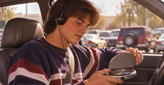 Teenager in a striped sweater sits in a car, wearing headphones and placing a CD into a portable CD player. Parked cars and trees are visible outside the window.