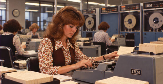 A woman in 1970s attire types on an IBM keypunch machine in an office, surrounded by other women working and large computers in the background.