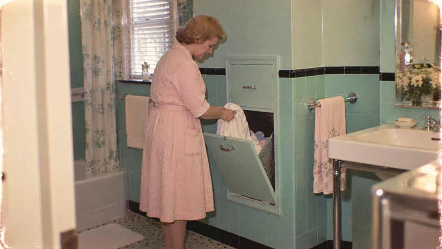 A woman in a pink robe places laundry into a built-in wall hamper in a teal-tiled bathroom.