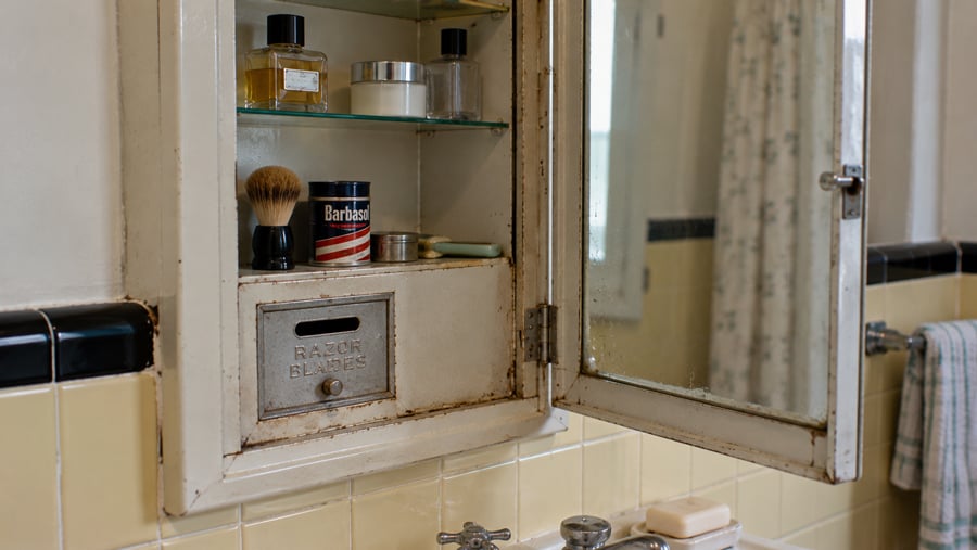 Open medicine cabinet above a sink containing glass bottles, a shaving brush, and a container of Barbasol, with a built-in metal slot labeled “Razor Blades.”