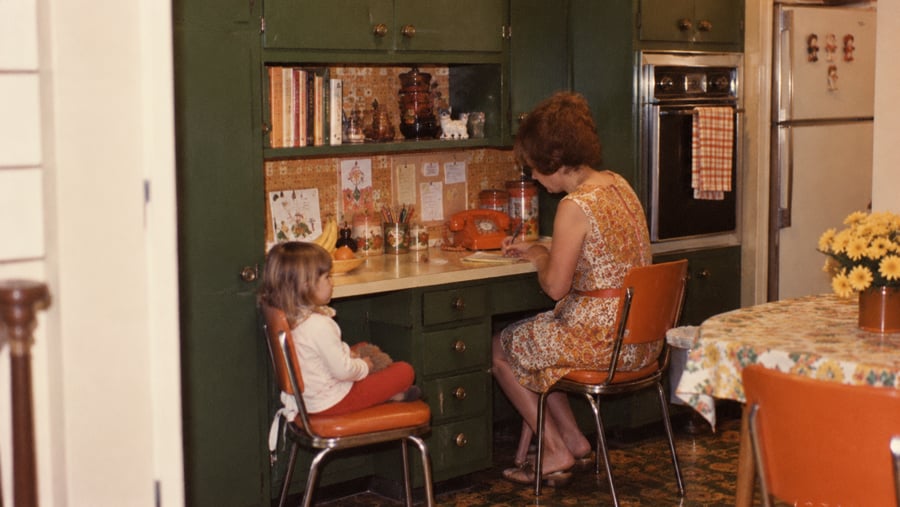 A woman sits at a kitchen desk writing, while a young child sits in a chair next to her.