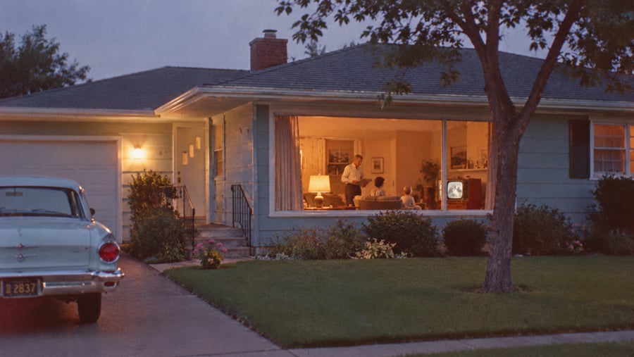 A vintage car parked in the driveway of a suburban house, with a family visible through the large living room window at twilight.