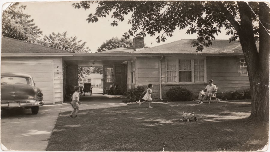 A vintage black and white photo shows a man sitting in a lawn chair under a tree while a young boy and girl play in the yard of a suburban house.