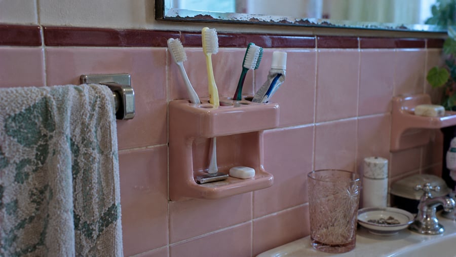 A wall-mounted pink ceramic toothbrush holder containing three toothbrushes and a tube of toothpaste, attached to a pink-tiled bathroom wall.