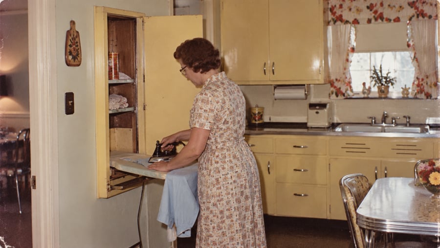A woman in a patterned dress irons a shirt on a fold-out board attached to a yellow kitchen cabinet.