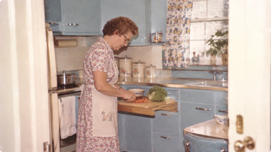 A woman in a floral dress and apron slices tomatoes on a cutting board in a vintage blue kitchen.