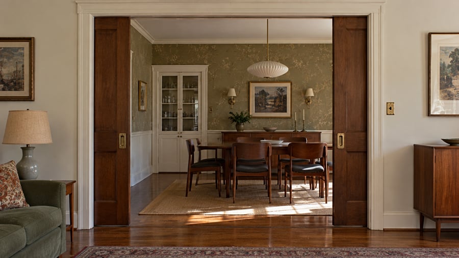 View from a living room through open wooden pocket doors into a dining room with a table, six chairs, and floral wallpaper.