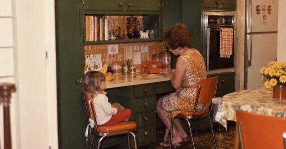A woman and a young child sit at a green built-in kitchen desk, with the woman writing in a notebook while the child watches.