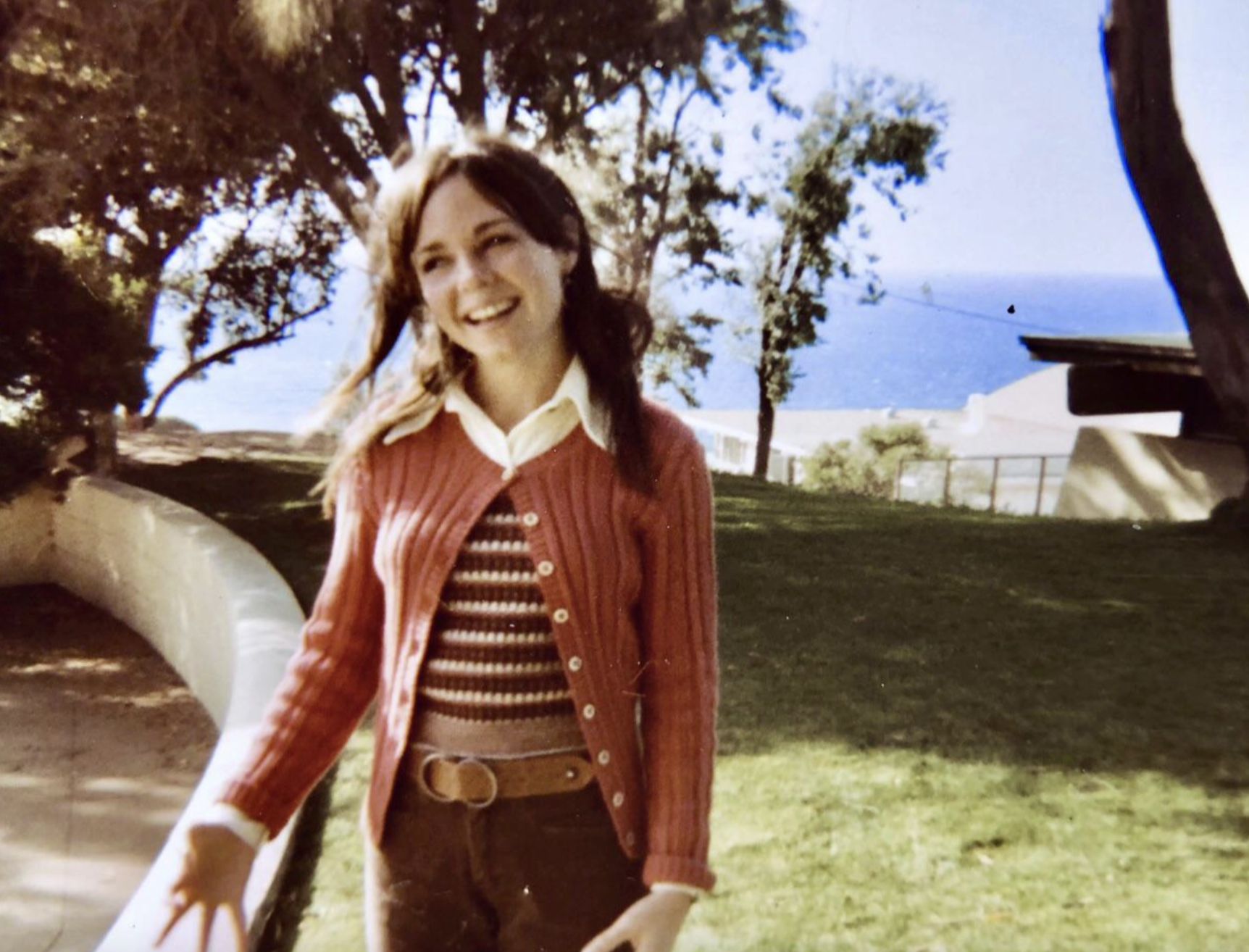 A smiling young woman in a striped shirt and pink cardigan stands in a park overlooking the ocean.