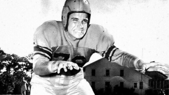 A vintage black-and-white photo of a football player in a leather helmet and jersey, crouched in a defensive stance.
