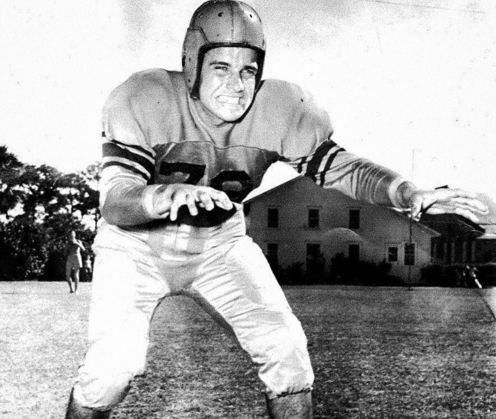 A vintage black and white photo of a football player in a leather helmet and uniform crouching in a defensive stance on a field.