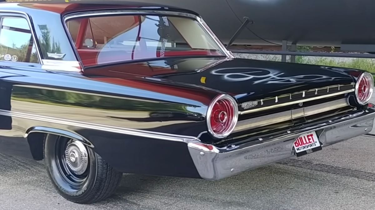 A black classic car with red circular tail lights and chrome trim is parked outdoors. The trunk has white graffiti, and the license plate reads “BULLET MOTORSPORTS.”