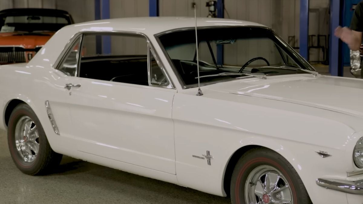 A classic white Ford Mustang coupe is parked indoors, with chrome wheels and the Mustang emblem on the side. Another vintage car in orange is partially visible in the background.