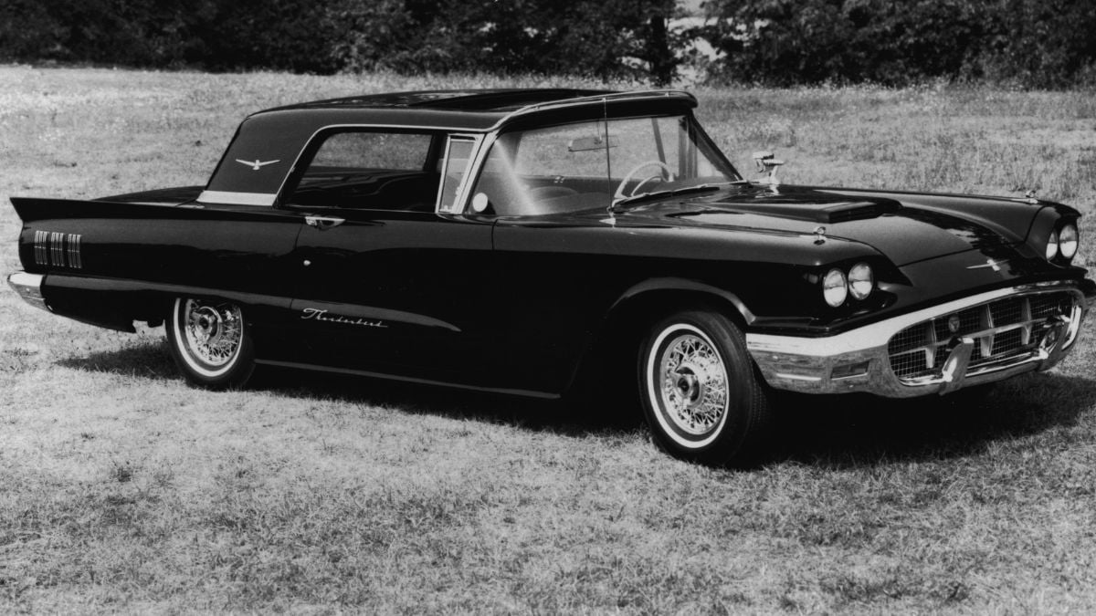 Black and white photo of a vintage Ford Thunderbird parked on grass, featuring a sleek design, white-wall tires, and a hardtop roof with a bird emblem. Trees and shrubs are visible in the background.