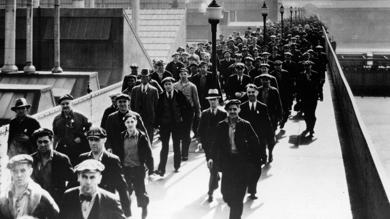 A large group of men in work clothes and hats walk together on a wide sidewalk or bridge, suggesting a historical industrial or factory setting. Black and white photo.