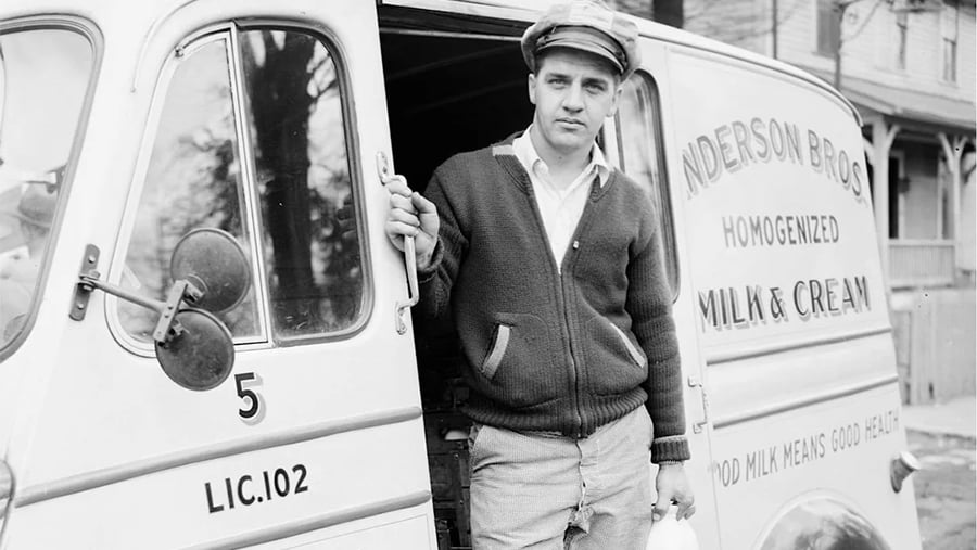 A young man in a cap and cardigan stands in the open doorway of an Anderson Bros. Milk & Cream delivery truck.