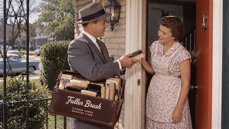 A Fuller Brush salesman in a suit and hat shows a brush to a woman standing in her doorway.