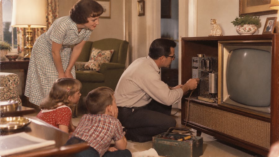 A man repairs the internal components of a large wooden cabinet television while a woman and two children watch.