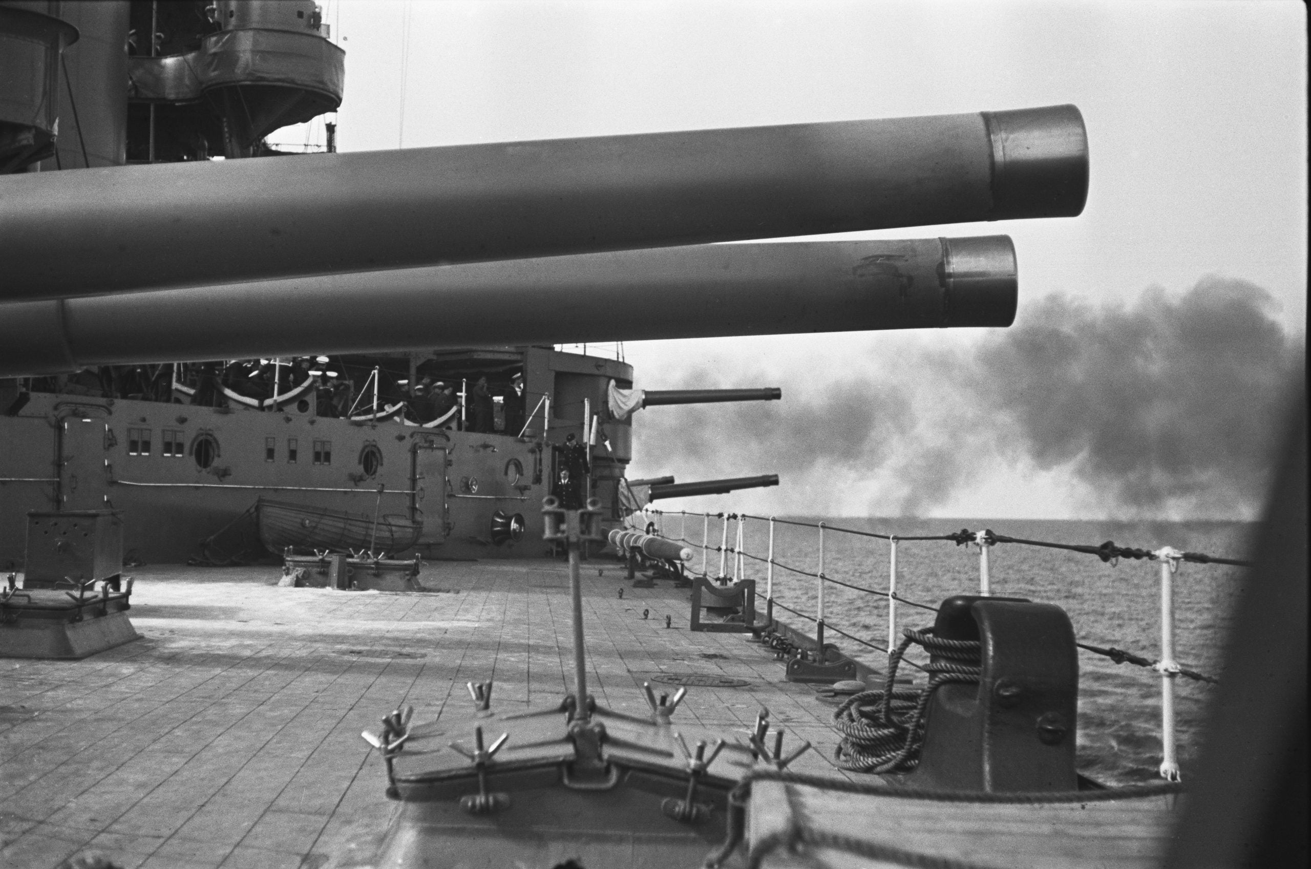 A battleship’s large guns aim toward the sea, with smoke rising on the horizon. The deck is visible with ropes and equipment.