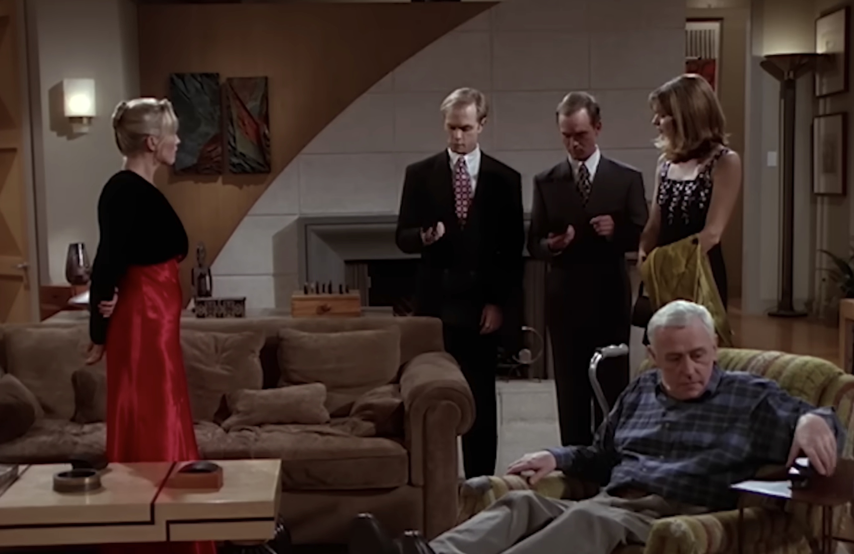 A woman in a red skirt and black top stands in a living room facing two men in suits and a woman holding a jacket, while an older man sits in a nearby armchair.